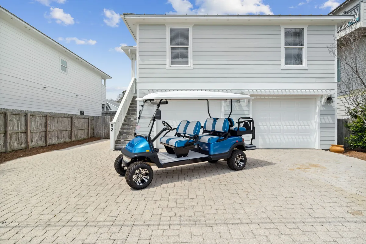 A close-up exterior shot featuring a blue and white 6-passenger street-legal golf cart parked in front of a two-car garage. The home’s light gray siding and exterior staircase complete the scene, suggesting easy transportation to local attractions and nearby beaches.