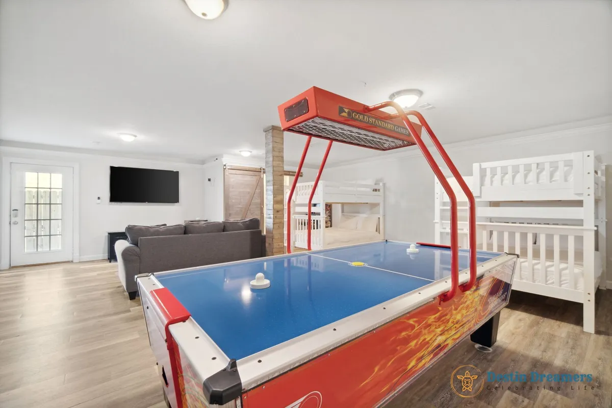 The scene captures a bustling entertainment space with an air hockey table in the foreground, white bunk beds along the side, a large sectional sofa facing a wall-mounted flat screen TV, and a rustic wooden barn door. The ambiance is playful, relaxed, and welcoming for groups or families.