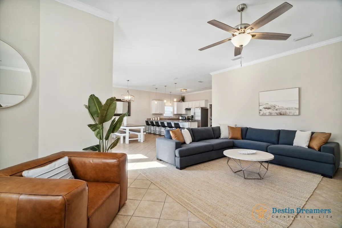 A spacious, open-concept interior featuring a large navy blue sectional sofa with accent pillows, a round coffee table, a brown leather armchair, and a view into the open kitchen with a breakfast bar and dining area. Natural light fills the space, emphasizing the comfortable and inviting atmosphere.