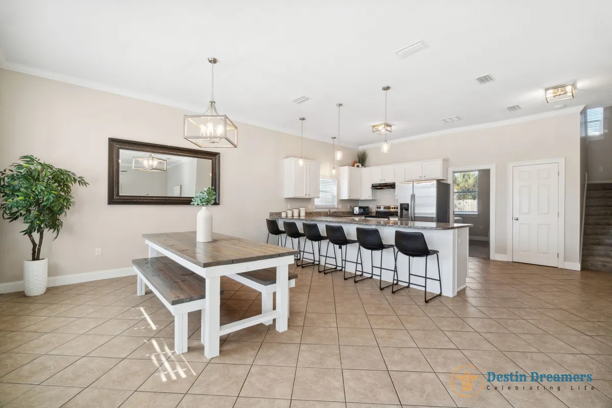 A bright, communal kitchen and dining space with a large island that seats six, a wooden dining table with bench seating, stainless steel kitchen appliances, and a large mirror on the wall. The layout encourages group cooking, dining, and socializing.