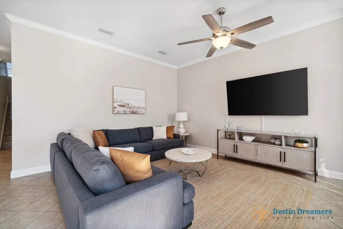 A spacious, inviting living area features two dark gray sofas with plush cushions, a large flat-screen TV mounted on the wall, a round coffee table, and a ceiling fan. The setting is ideal for watching movies, sports, or simply relaxing with family or friends after a day of activities.
