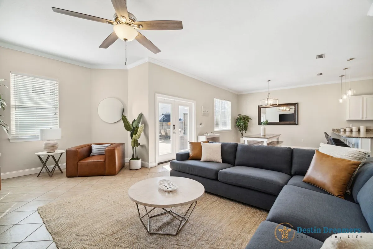 A cozy, open-plan living and dining area with a large dark gray sectional sofa, accent pillows, a round coffee table, a brown leather armchair, and a dining area with bench seating and pendant lighting. The setting is designed for group connection and relaxation.