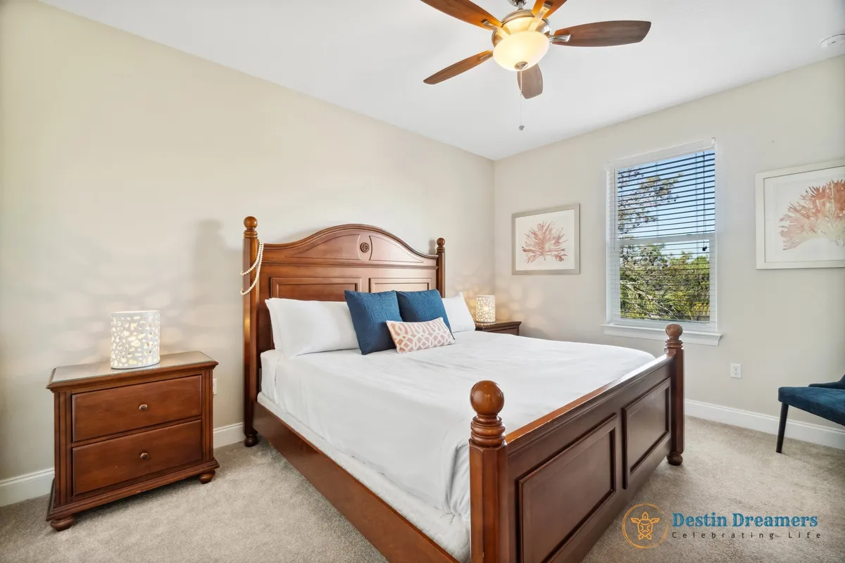 A queen bedroom with a classic wooden bed, white plush linens, matching nightstands with decorative lamps, and a window letting in natural light with views of greenery. Framed coral artwork adds a touch of coastal tranquility.