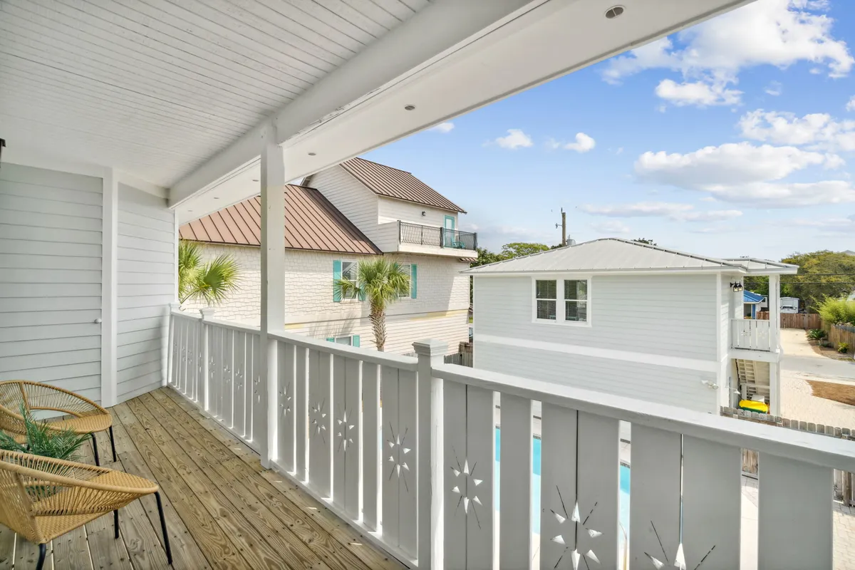 A covered balcony with rustic wooden flooring and decorative white railing, natural wood chairs for relaxation, and a view of the carriage house and neighboring buildings framed by palm trees. The mood is peaceful and welcoming, hinting at tranquil moments after days of adventure.