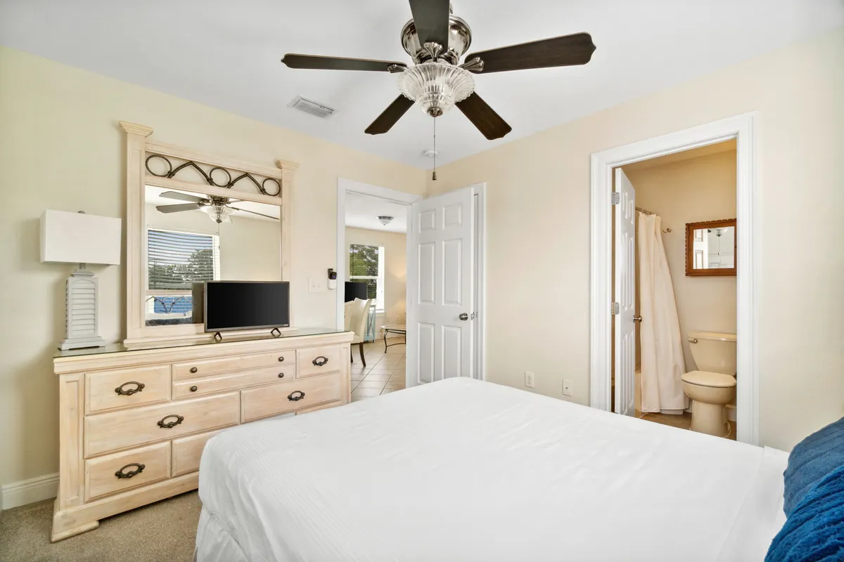 A spacious bedroom featuring a king size bed with crisp white linens, a dresser with mirror and small TV, and a ceiling fan. An en-suite bathroom entrance is visible, enhancing the sense of privacy and luxury. The room is bright and inviting, ideal for restful sleep.