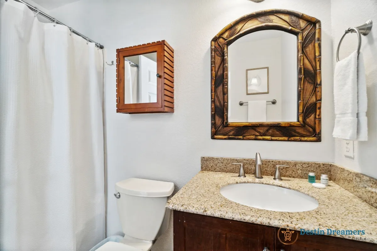 A stylish bathroom with an ornate wooden framed mirror above a granite countertop, a white shower curtain, and a toilet. The space is fresh, bright, and inviting, offering a spa-like feel for personal routines.