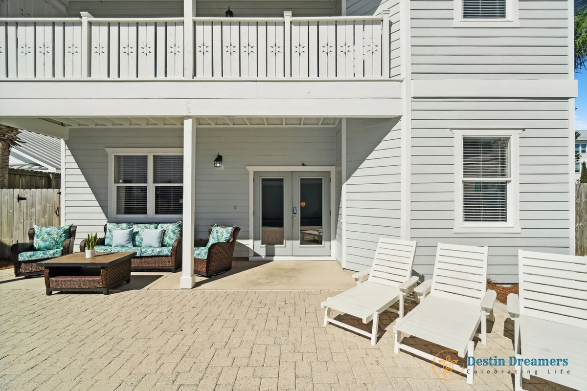 A private outdoor courtyard featuring comfortable wicker seating with turquoise cushions, three white lounge chairs, and potted plants on a coffee table. The inviting white double doors lead to a carriage house queen bedroom, all framed by a peaceful, sunlit setting.
