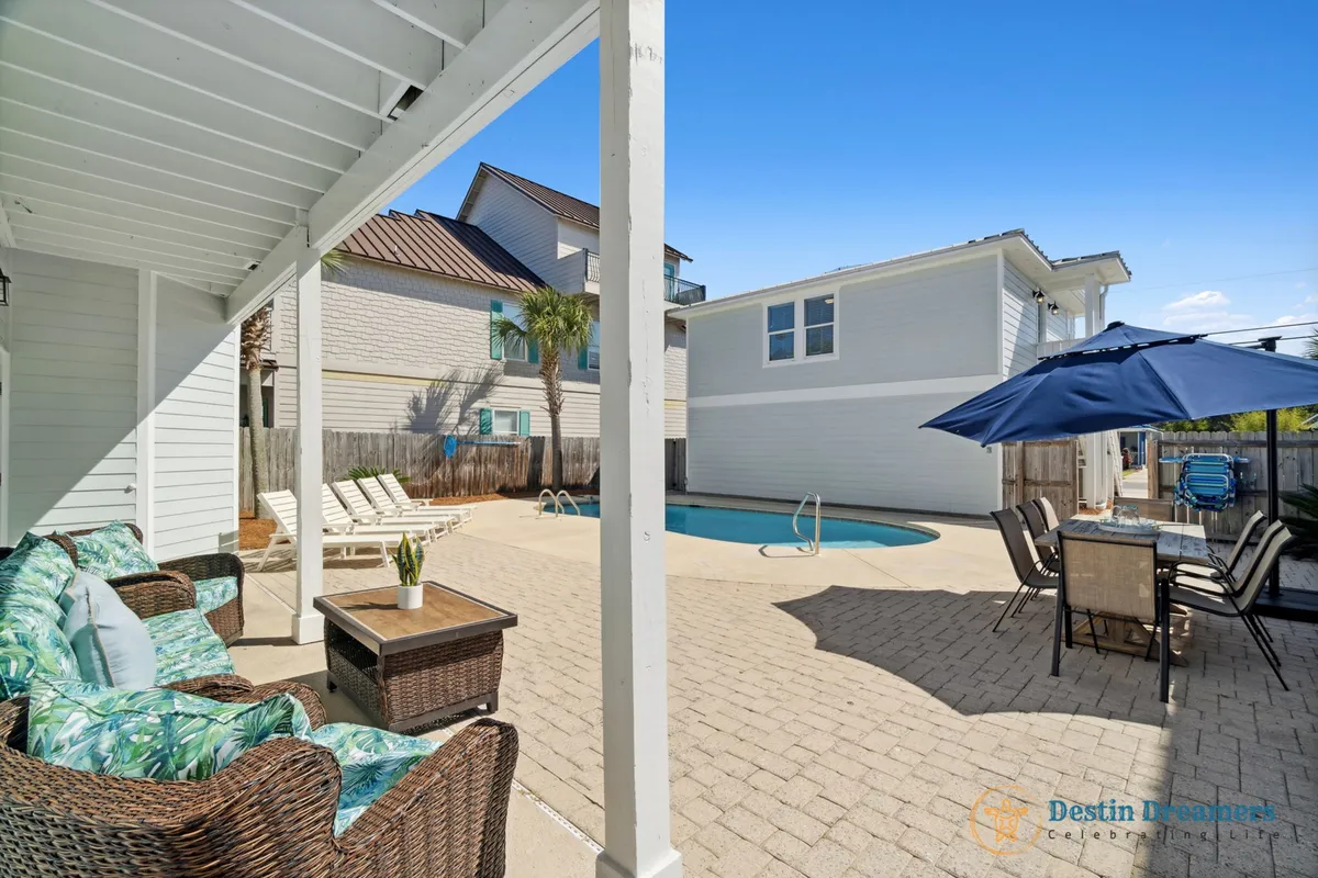 The scene centers on a serene pool area with wicker outdoor seating featuring tropical cushions and an outdoor dining table shaded by a blue umbrella. The carriage house is visible, and the layout encourages both relaxation and social connection. The area feels intimate and comfortable, ideal for small groups or families.