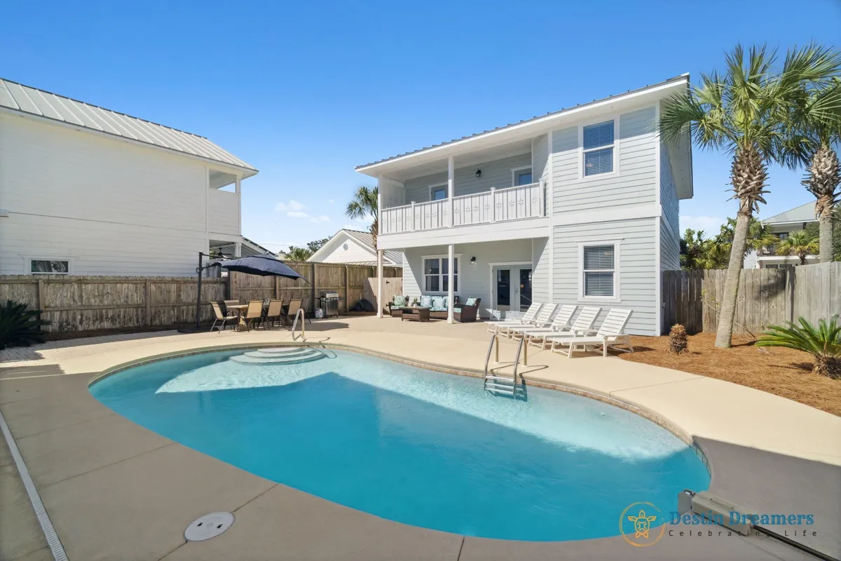 This image showcases a private heated pool with an attached spa, surrounded by lounge chairs and an outdoor dining area with an umbrella. The two-story carriage house with a balcony provides a picturesque backdrop, and the entire area is enclosed for privacy and relaxation. The lush greenery and thoughtful layout evoke a resort-like atmosphere, perfect for leisure and group gatherings.