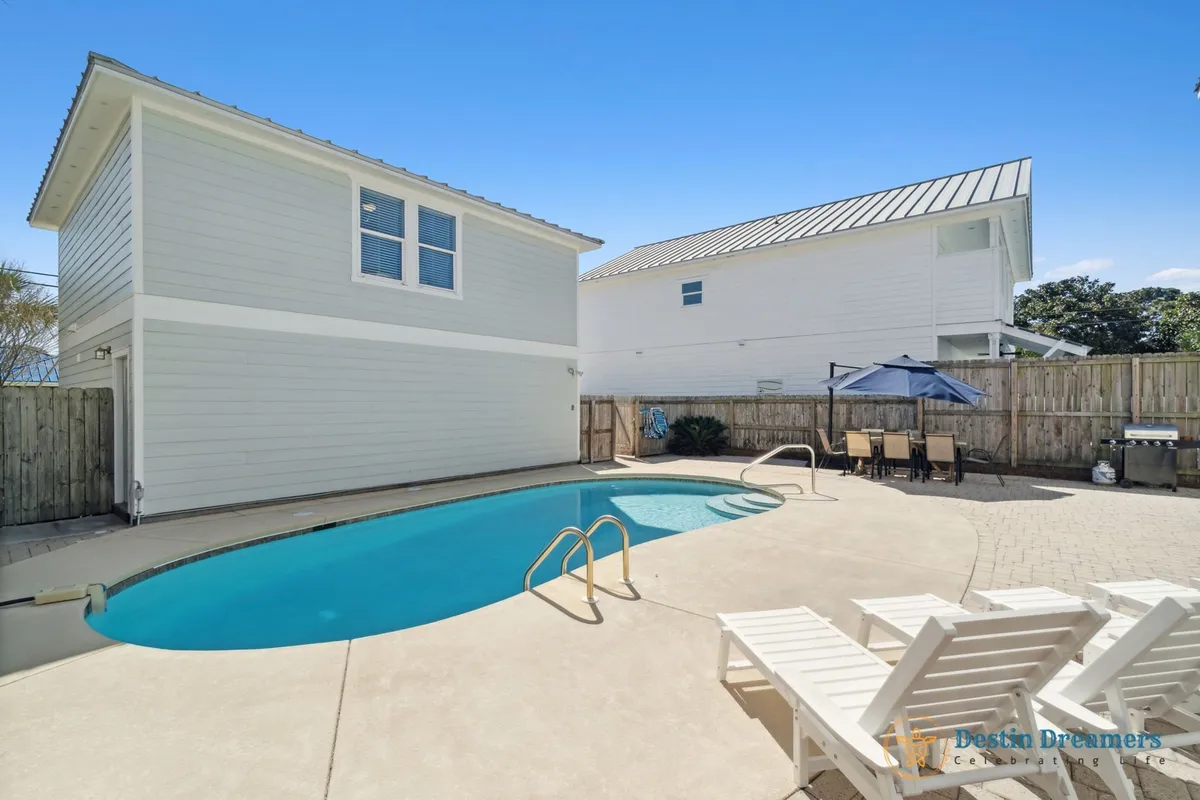 A private heated swimming pool invites relaxation, surrounded by white lounge chairs, an outdoor dining table with an umbrella, and a barbecue grill. The secluded setting is perfect for family cookouts, poolside lounging, and sunny afternoons spent in comfort and privacy.
