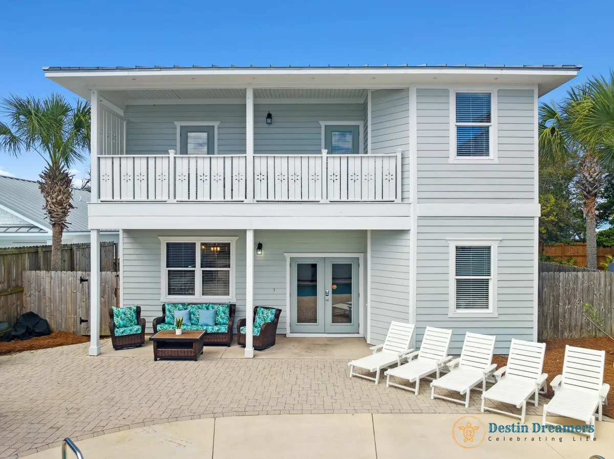 A second-floor balcony with decorative railing overlooks a row of white lounge chairs beside a pool. The space features comfortable seating with tropical cushions and double glass doors leading inside, evoking a relaxed, coastal vacation atmosphere perfect for both people and pets.