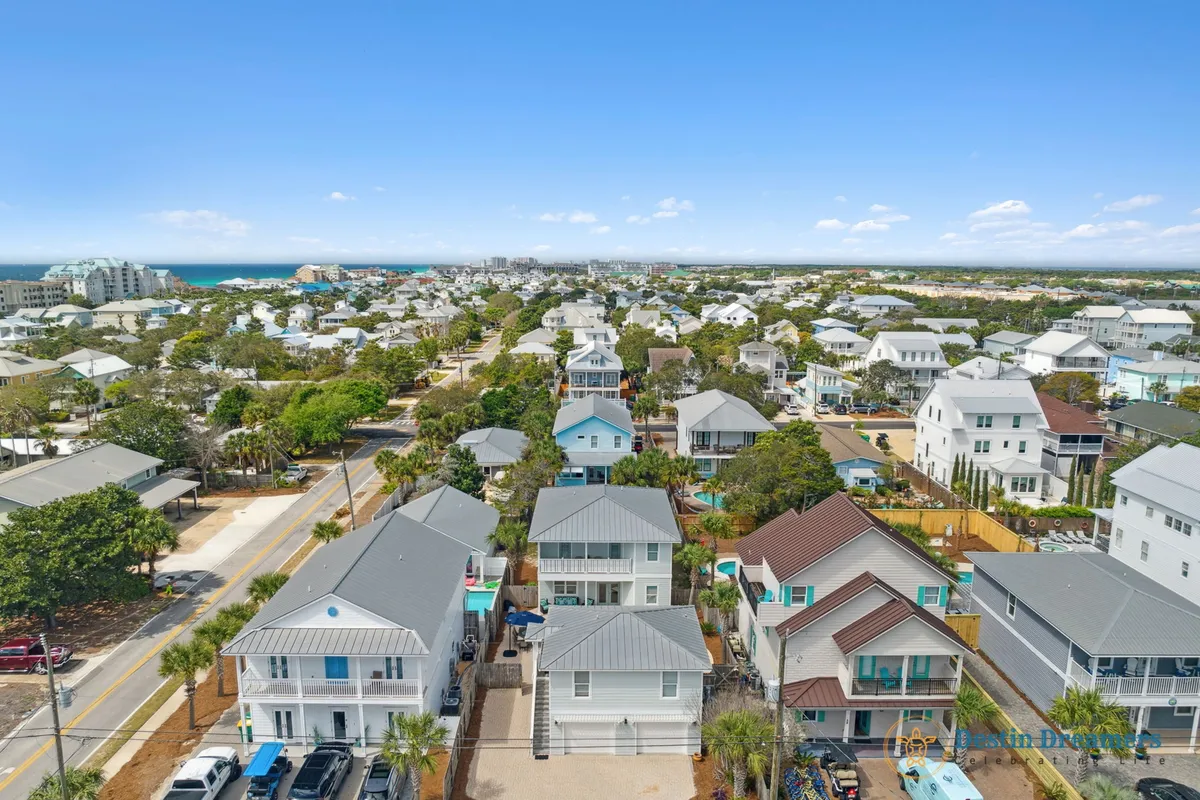 A scenic neighborhood view with rows of beach-style homes, metal roofs, tree-lined streets, balconies, and distant ocean views. The image evokes a relaxed, inviting coastal lifestyle with easy access to both the beach and local amenities.