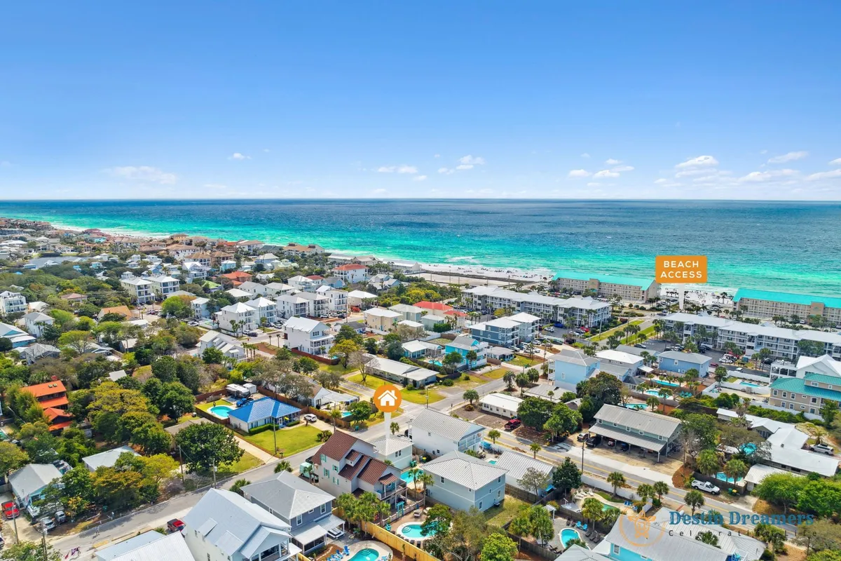 This exterior shot captures the turquoise ocean with visible beach access, residential houses with pools, and both the main house and carriage house with a private pool. The setting is lively, showing an inviting and relaxing coastal community ideal for group vacations and seasonal activities.