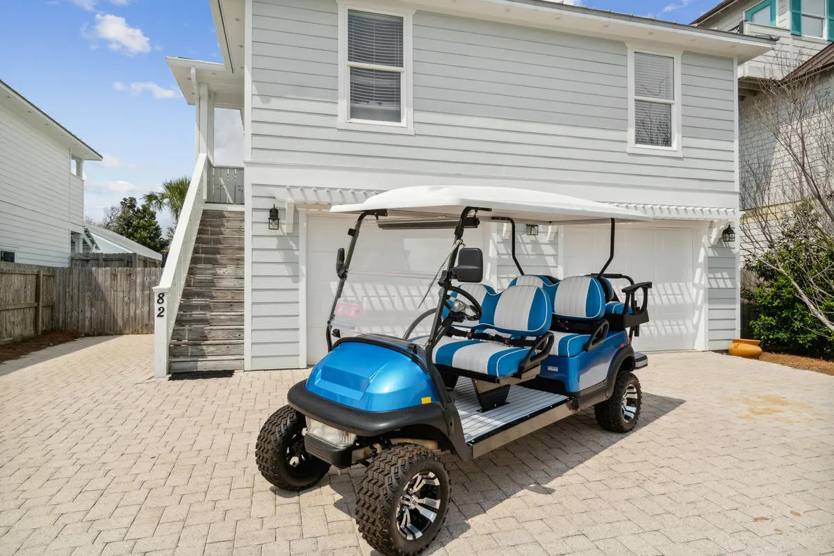 A blue and white 6-passenger street-legal golf cart stands ready in front of a detached garage, with the light gray-sided house and exterior stairs in the background. The scene radiates a laid-back, vacation-ready mood and signals easy group outings to the beach.